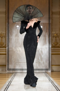 Model on a runway wearing a black glittery gown with a large sheer black fan held near her face.