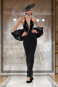 Model on a runway wearing a black glittery gown with a deep V neckline and dramatic oversized puff sleeves, paired with a wide-brimmed hat.