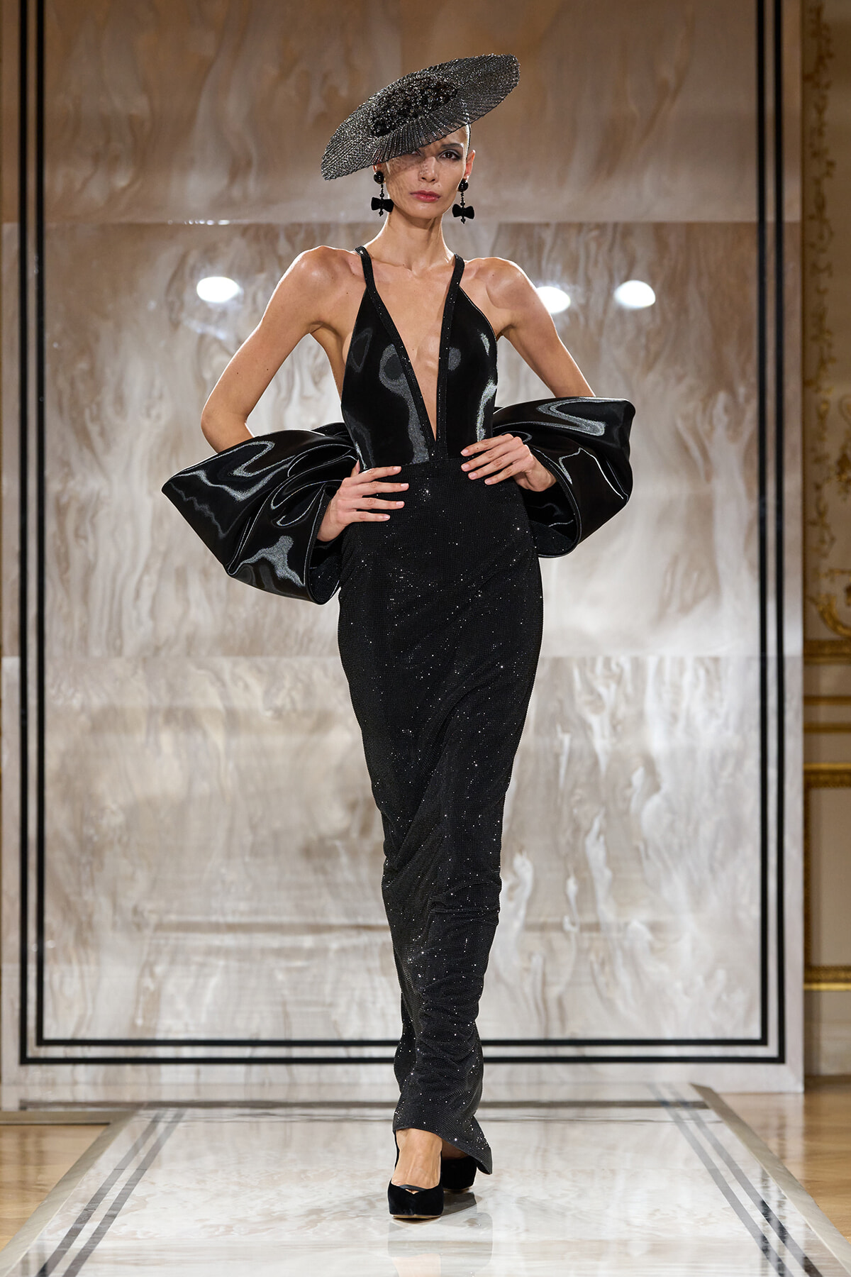 Model on a runway wearing a black glittery gown with a deep V neckline and dramatic oversized puff sleeves, paired with a wide-brimmed hat.