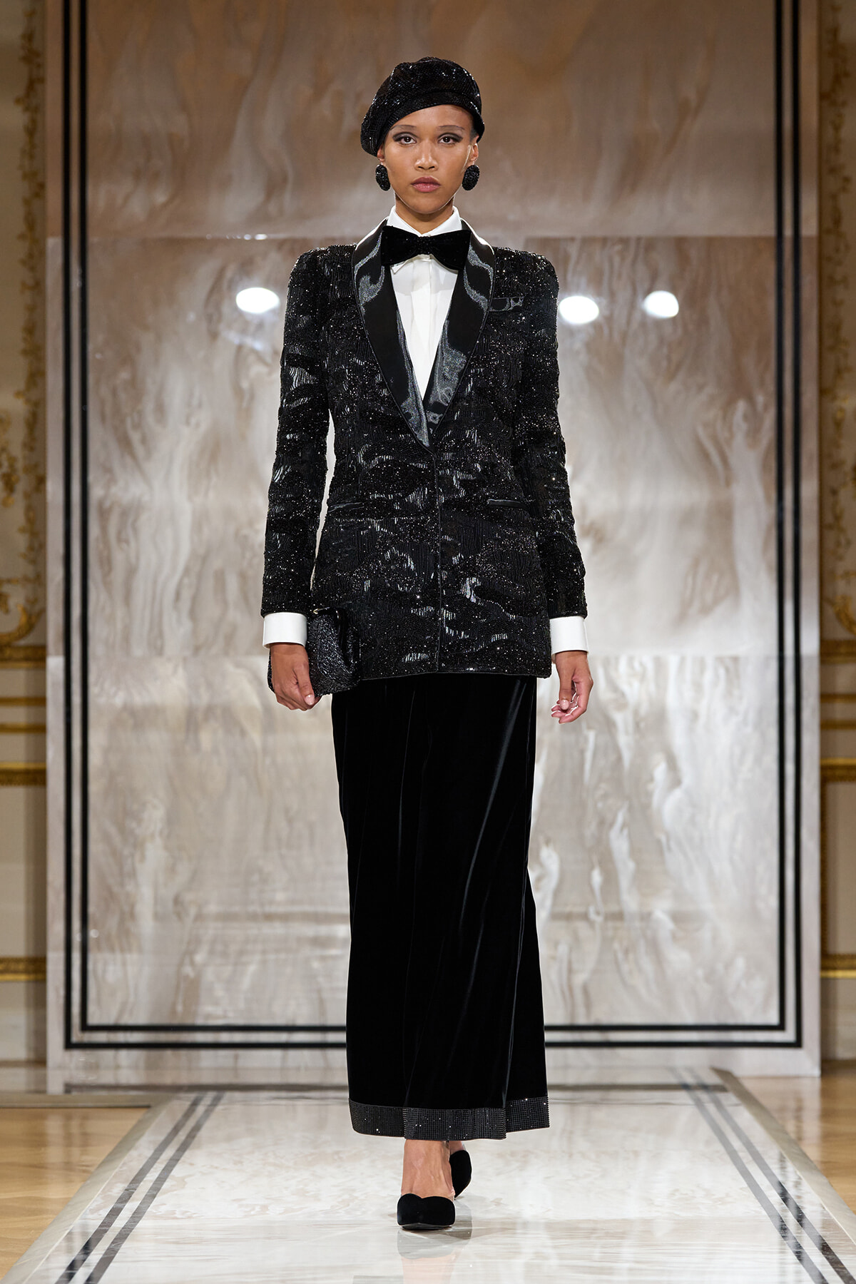 Runway look: a model in a black sequined blazer with satin lapels, white shirt, bow tie, and a long black velvet skirt with cuffed white shirt sleeves showing.