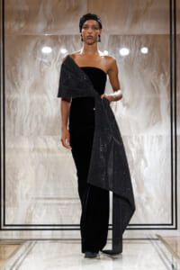 Model on a runway wearing a black velvet one-shoulder gown with a sparkling wrap draped over her arm and a matching headscarf.