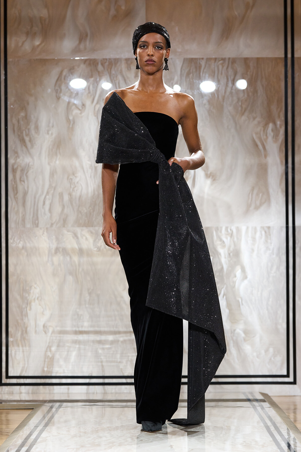 Model on a runway wearing a black velvet one-shoulder gown with a sparkling wrap draped over her arm and a matching headscarf.