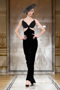 Model walking a runway in a black velvet gown with cutout sides and a beaded bralette-style top, paired with a large black textured hat on a marble catwalk.