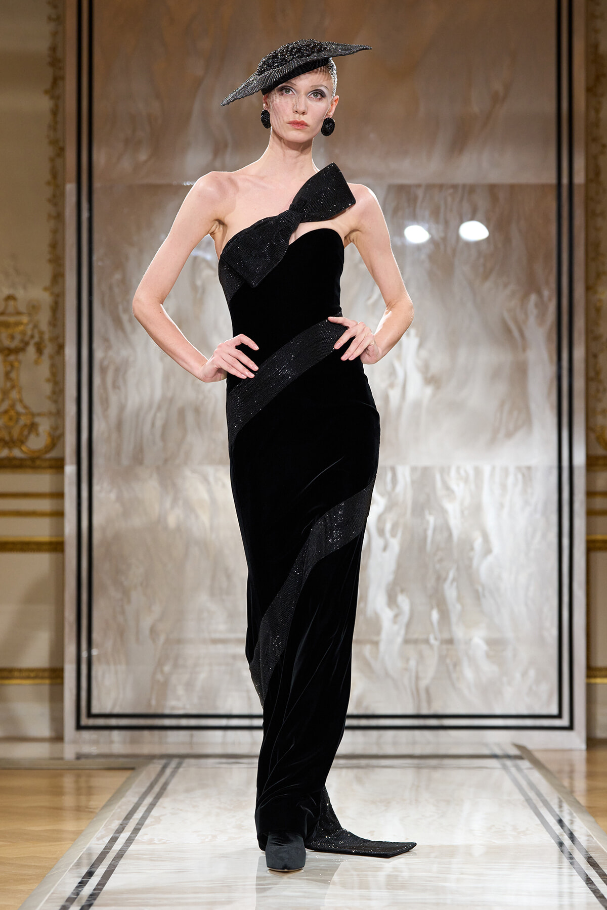 Runway model in a strapless black velvet gown with a glittery bow, diagonal sash, and matching decorative hat.