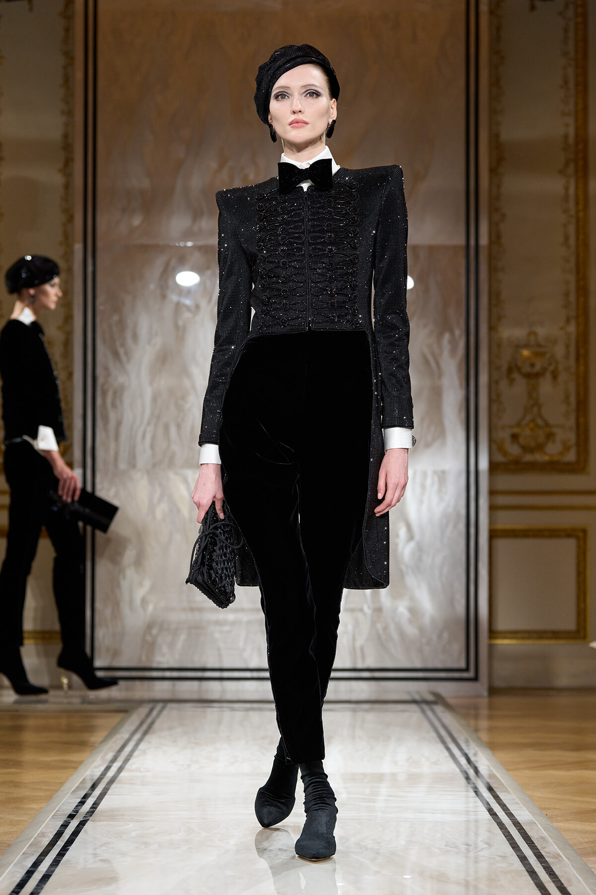 Runway model in a black sequined tailored jacket with white cuffs, bow tie, velvet pants, and a black head wrap, carrying a small black bag on a catwalk.