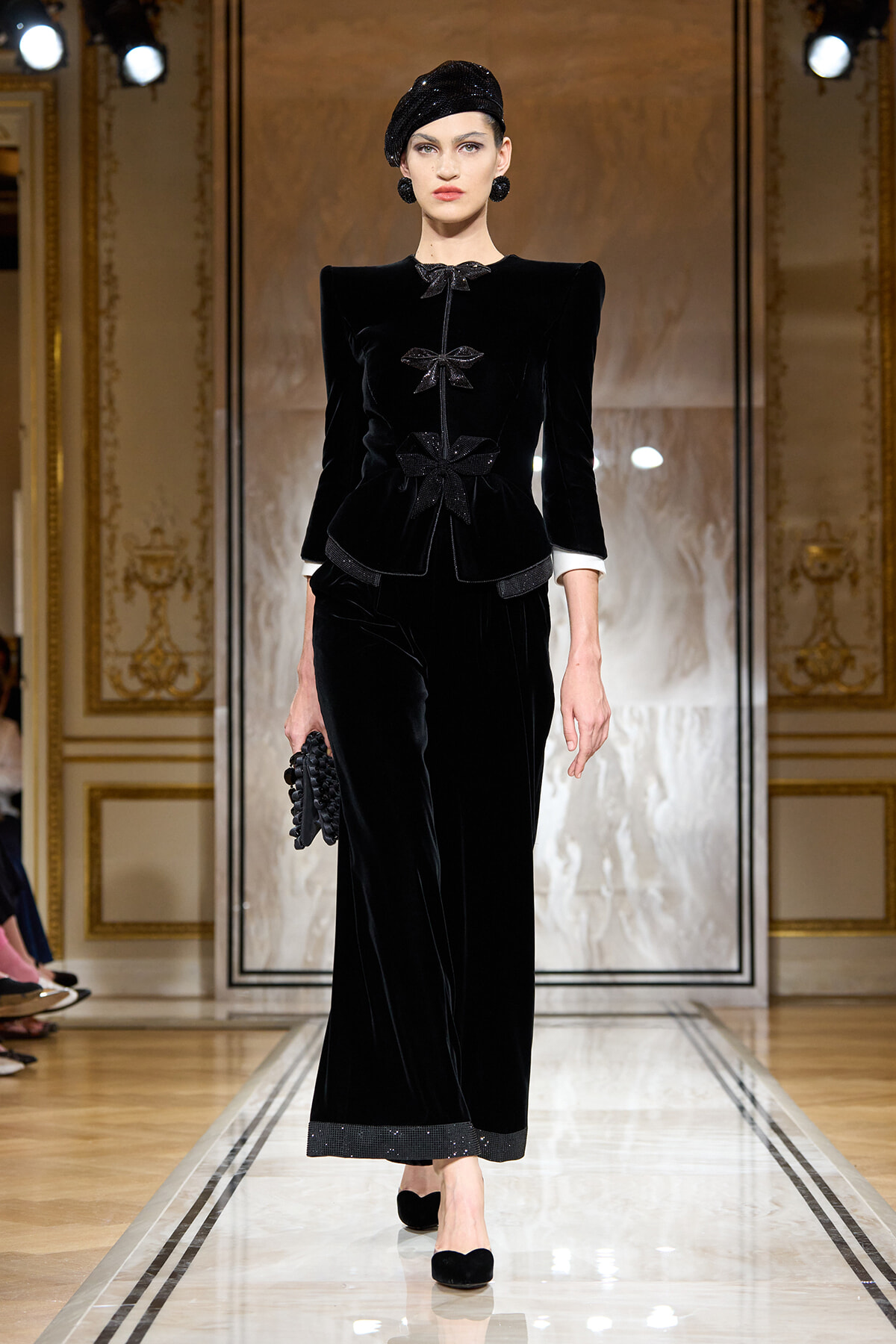 Model on a runway wearing a black velvet suit with bow embellishments, matching hat, and black accessories in a luxe gold-trimmed setting.
