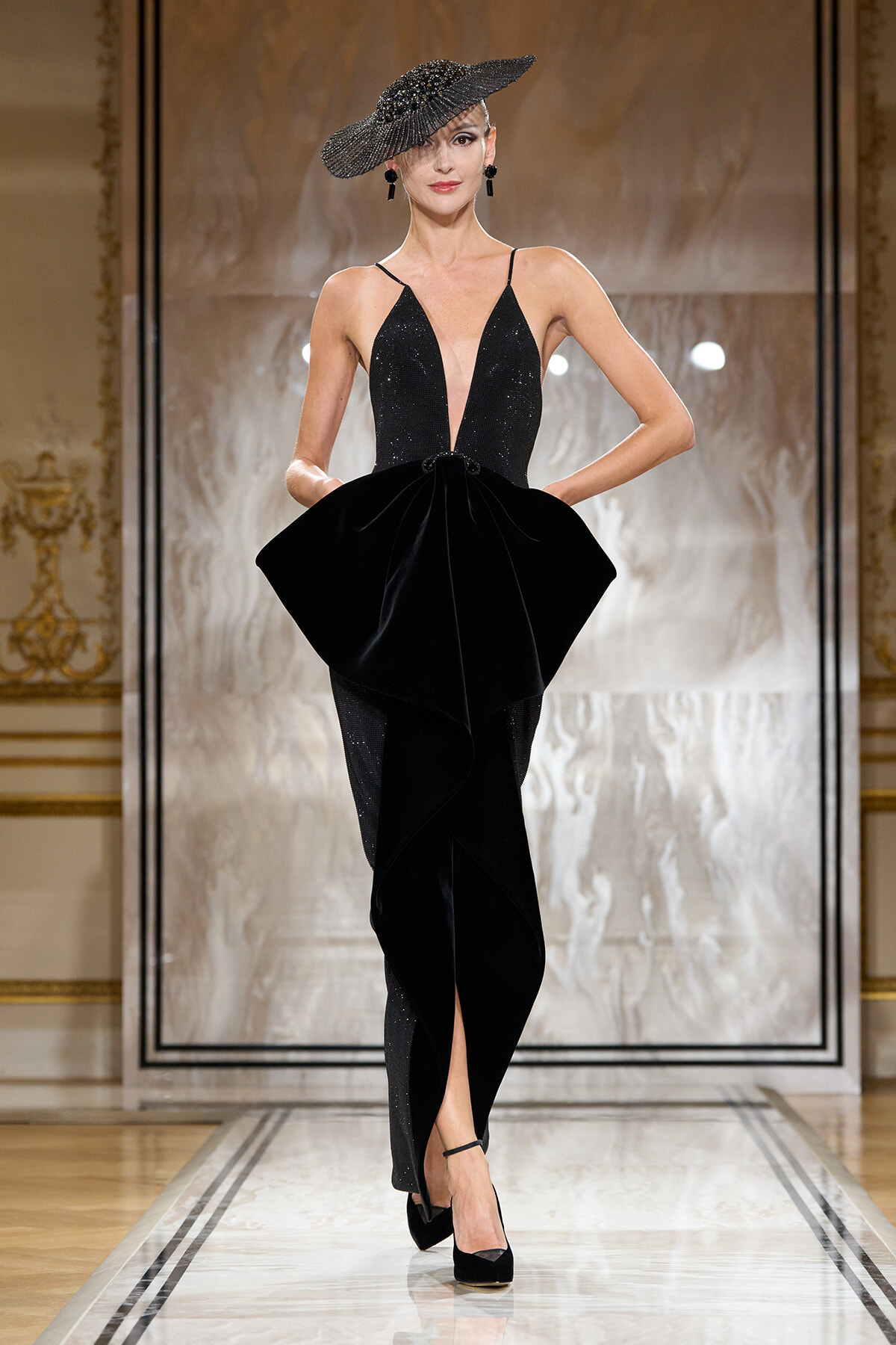 Runway model in a sleek black gown with a plunging neckline, dramatic sculpted peplum at the waist, and a glittering wide-brim hat.