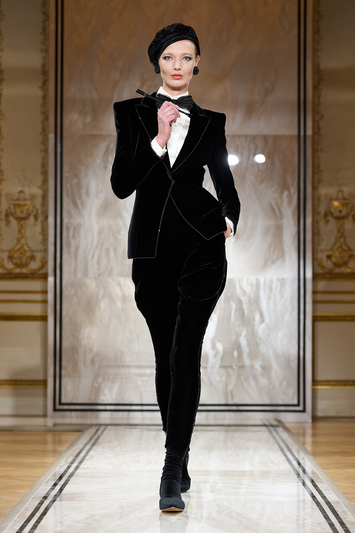 Female model on a runway wearing a black velvet tailored suit with white shirt, bow tie, and a black headwrap.
