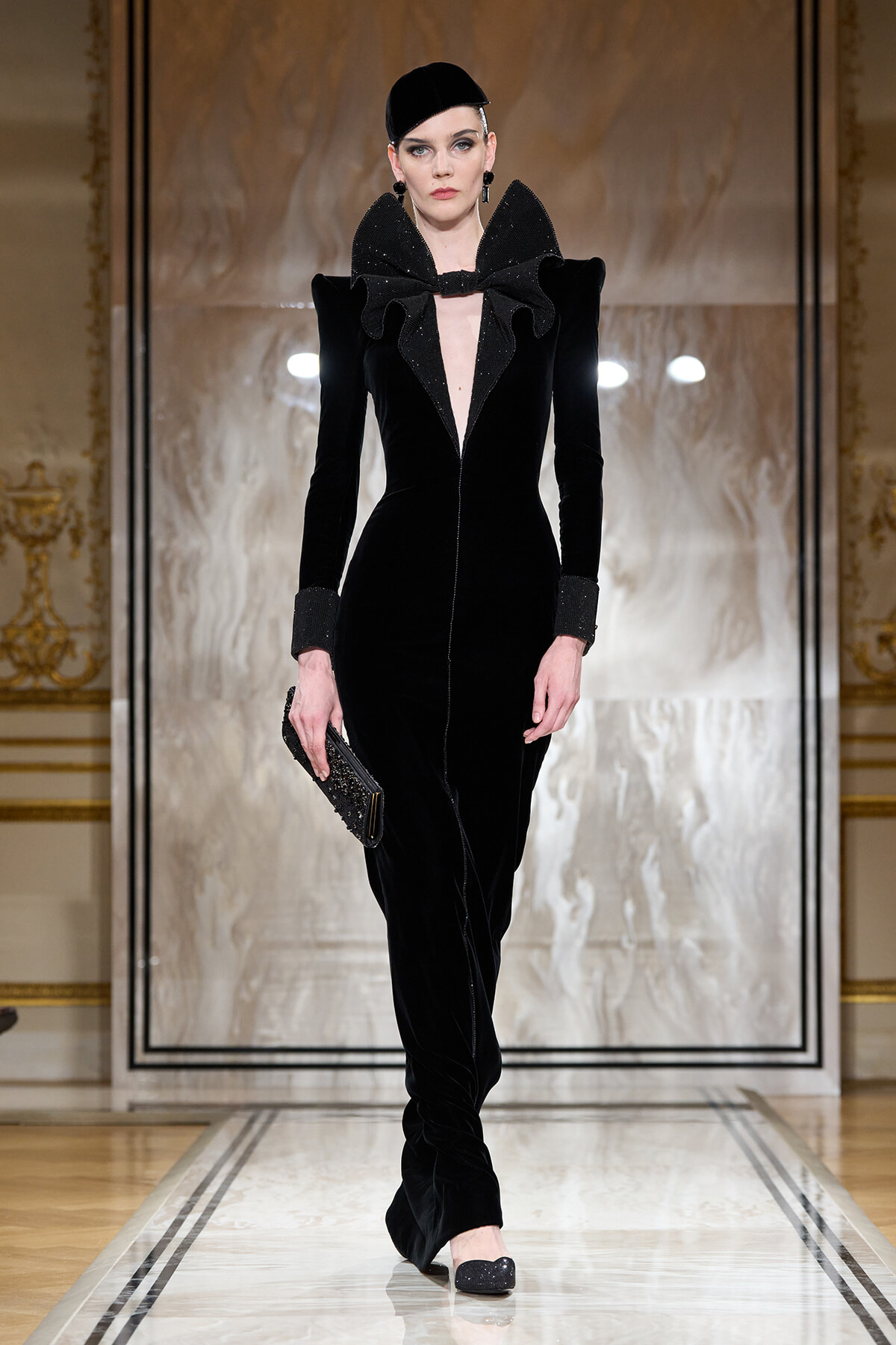 Model on a runway wearing a black velvet jumpsuit with a large ruffled collar, long sleeves, and a matching cap, carrying a glittery clutch.