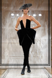 Model on a runway wearing a dramatic black strapless jumpsuit with a plunging V and oversized bow-like hips, paired with a metallic sculpted hat and dark socks/heels