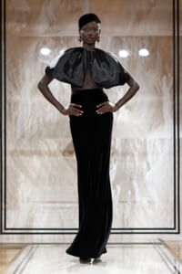 Model on a runway wearing a sleek black velvet column gown with a voluminous metallic capelet and a black cap, accessorized with bold earrings.