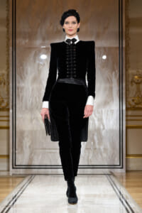 Model on a runway wearing a black velvet tailored suit with white shirt cuffs, bow tie, head wrap, and a black clutch.