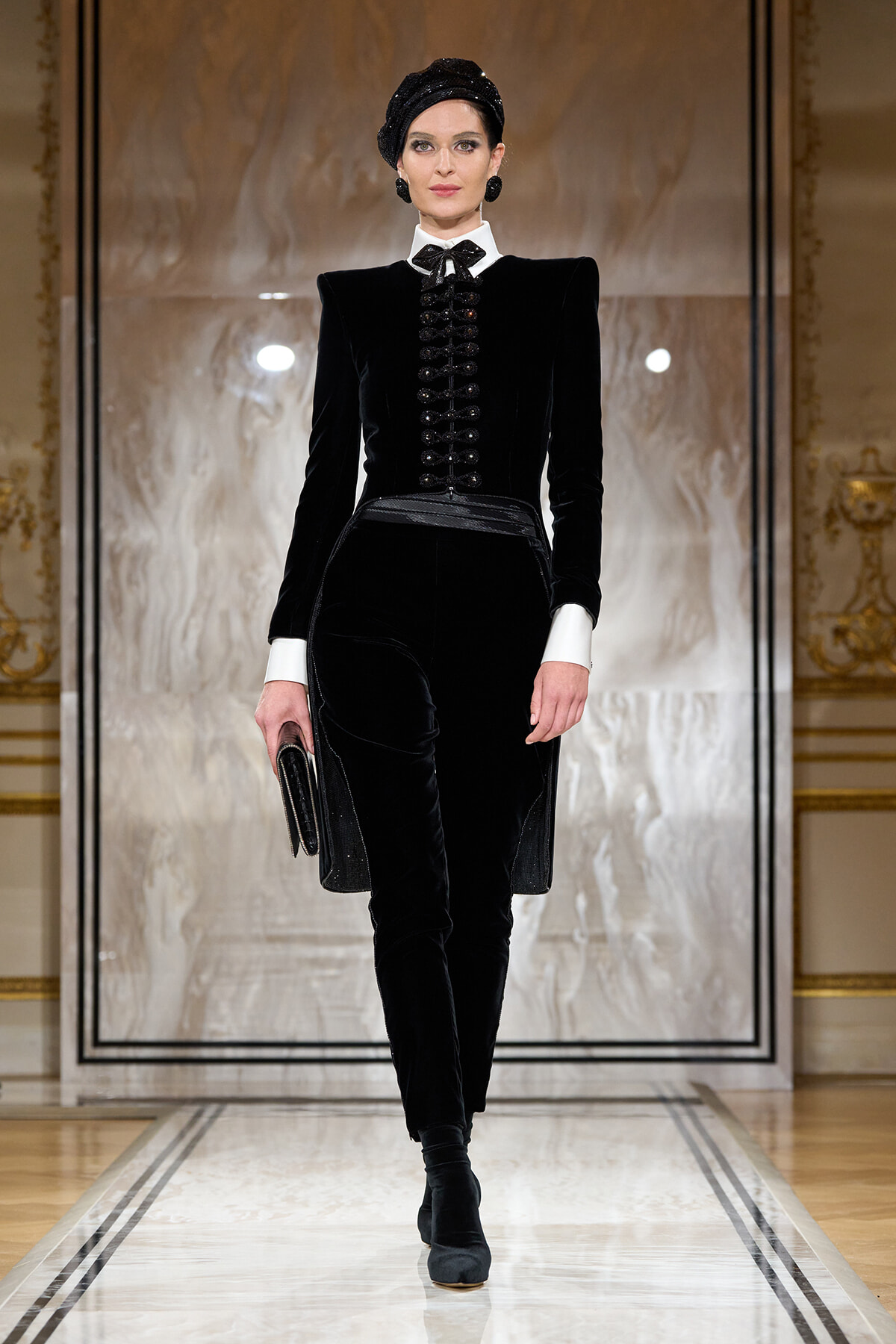 Model on a runway wearing a black velvet tailored suit with white shirt cuffs, bow tie, head wrap, and a black clutch.