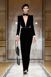 Model on a runway wearing a black velvet gown with a plunging neckline, white cuffs, a bow tie, and a small black tasselled handbag.
