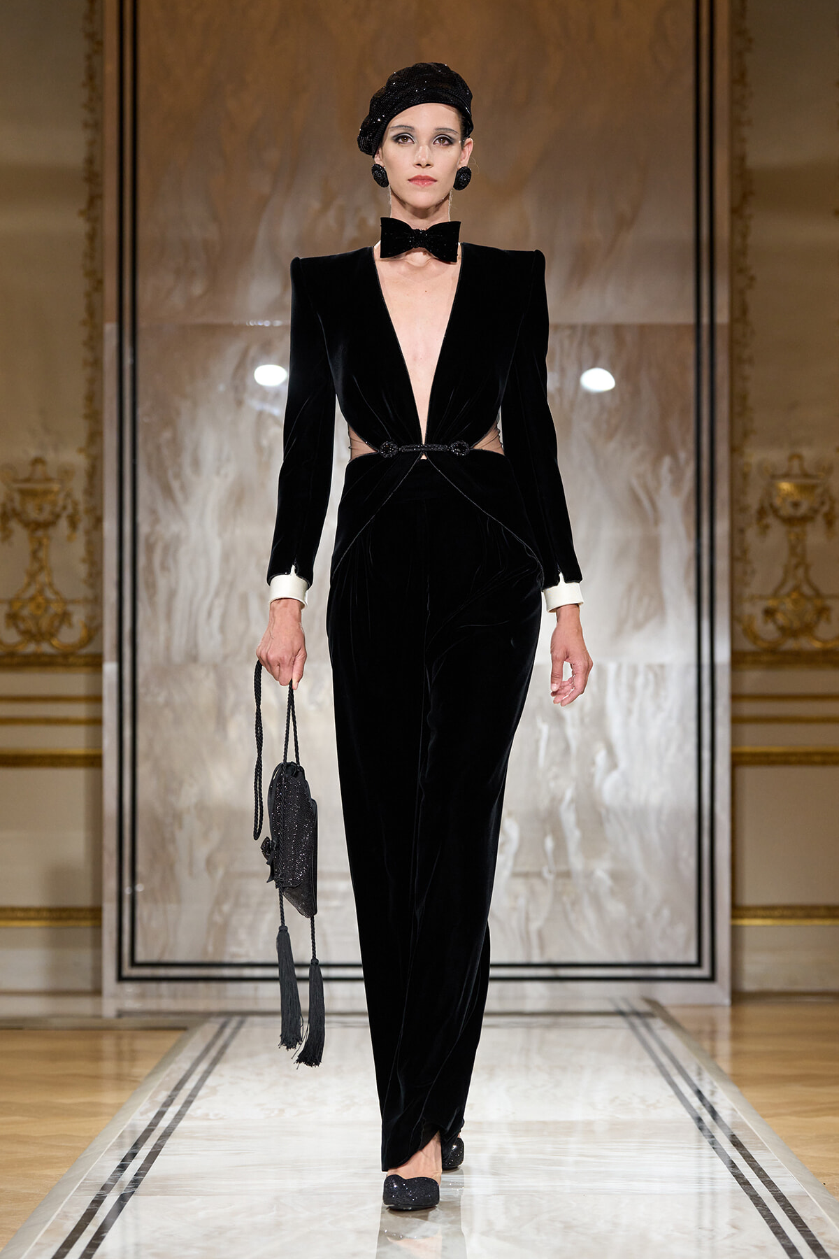 Model on a runway wearing a black velvet gown with a plunging neckline, white cuffs, a bow tie, and a small black tasselled handbag.