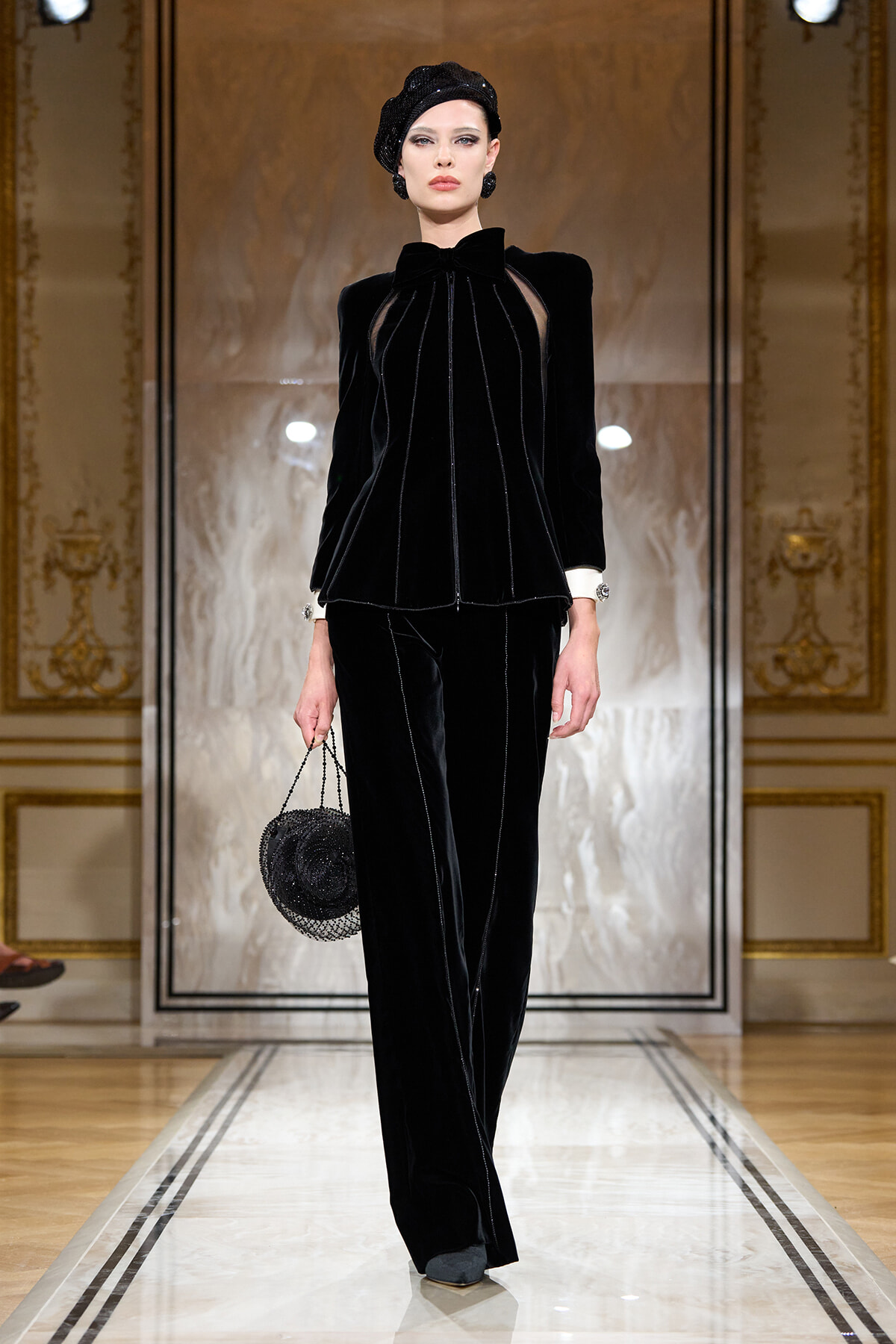 Model in a black velvet pantsuit with sheer inset details, matching beret, walking a runway in a gilded, ornate setting.