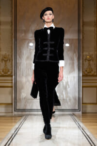 Model on a fashion runway wearing a black velvet suit with white shirt cuffs, bow tie, and a black beret, in a gilded setting.