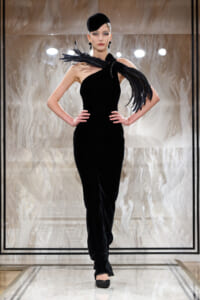 Model on runway wearing an elegant black one-shoulder velvet gown with feathered shoulder accessory and a small black hat.