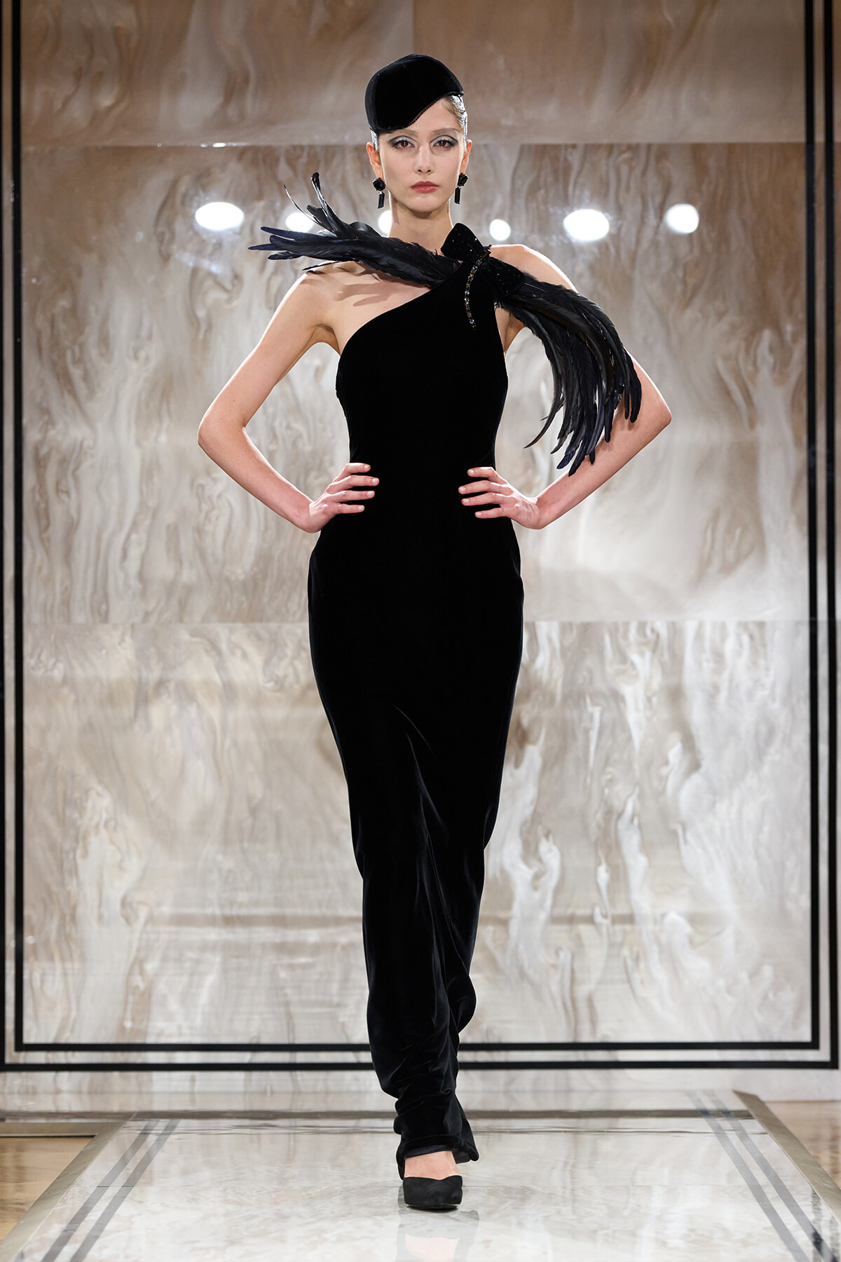 Model on runway wearing an elegant black one-shoulder velvet gown with feathered shoulder accessory and a small black hat.
