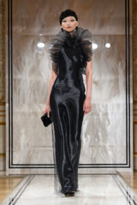 Fashion model walking the runway in a black, shimmery evening gown with dramatic sheer ruffled collar and matching headpiece, holding a small black clutch.