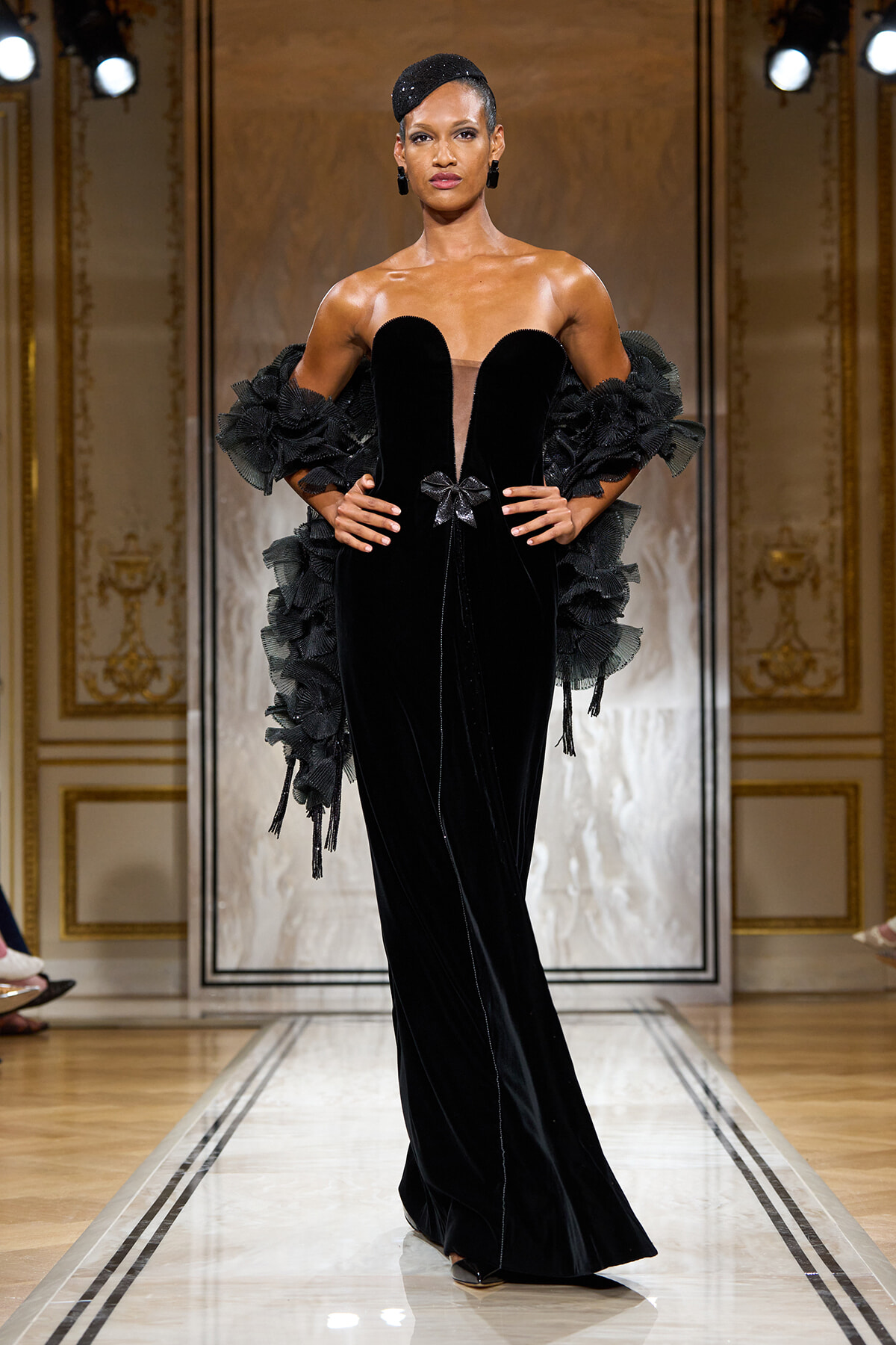Model on a runway wearing a floor-length black velvet gown with a sheer central panel, bow at the waist, and dramatic ruffled sleeves.