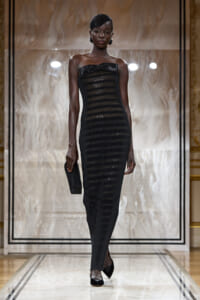 Model wearing a strapless black sequined striped gown with a bow at the bust, a matching headpiece, and a small black clutch on a fashion runway.