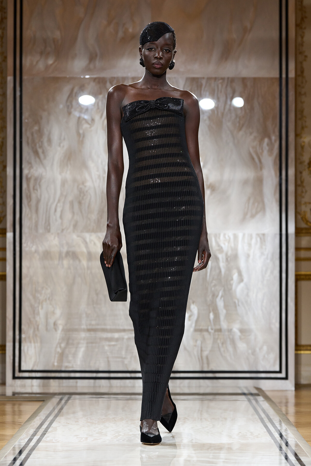 Model wearing a strapless black sequined striped gown with a bow at the bust, a matching headpiece, and a small black clutch on a fashion runway.