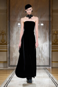Model on a glossy runway wearing a strapless black velvet gown, black bow choker, and matching hat in an ornate setting.