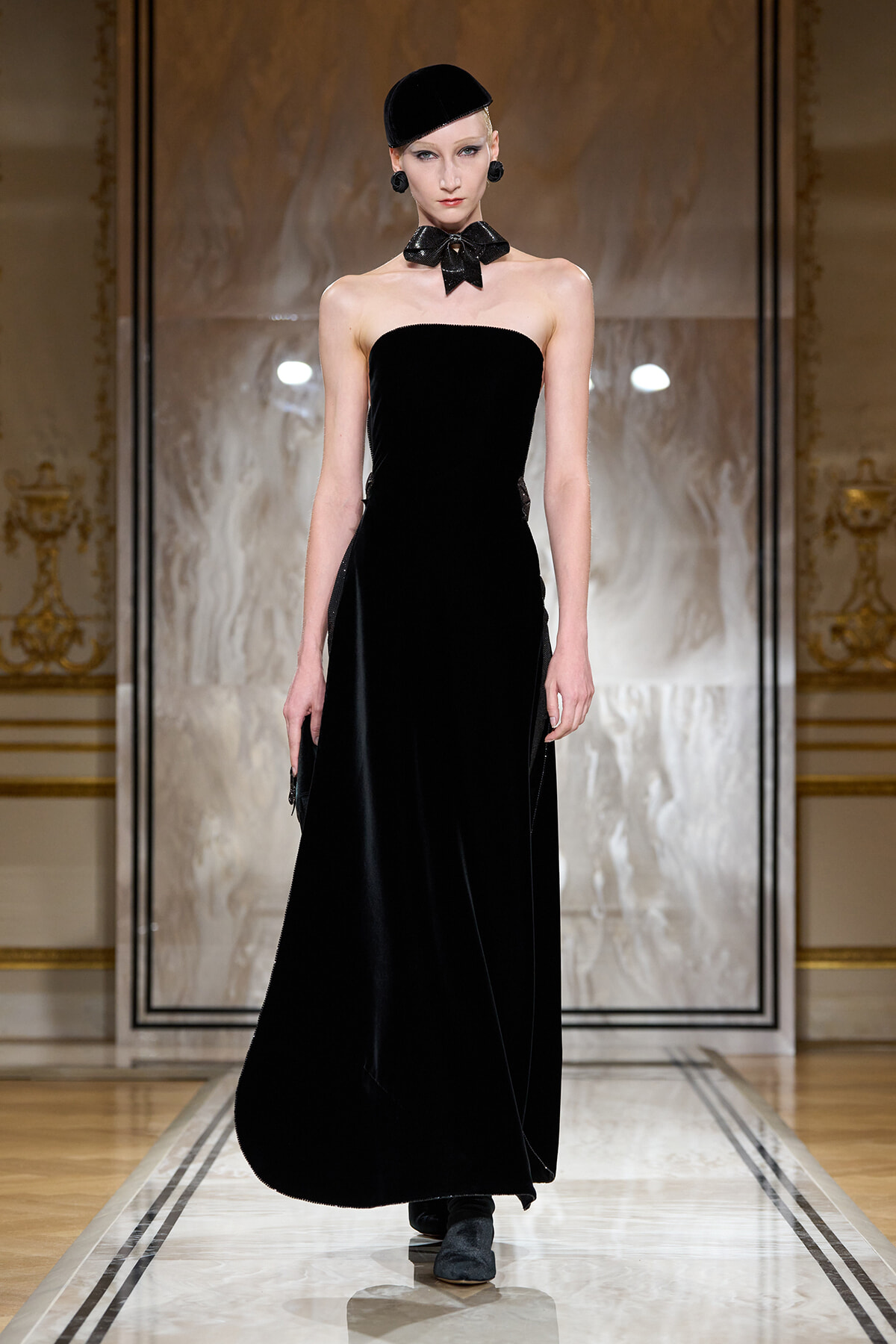 Model on a glossy runway wearing a strapless black velvet gown, black bow choker, and matching hat in an ornate setting.