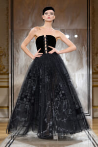 Model on a runway wearing a strapless black ball gown with a laced front bodice and a voluminous dotted tulle skirt, complemented by a black cap and statement earrings.