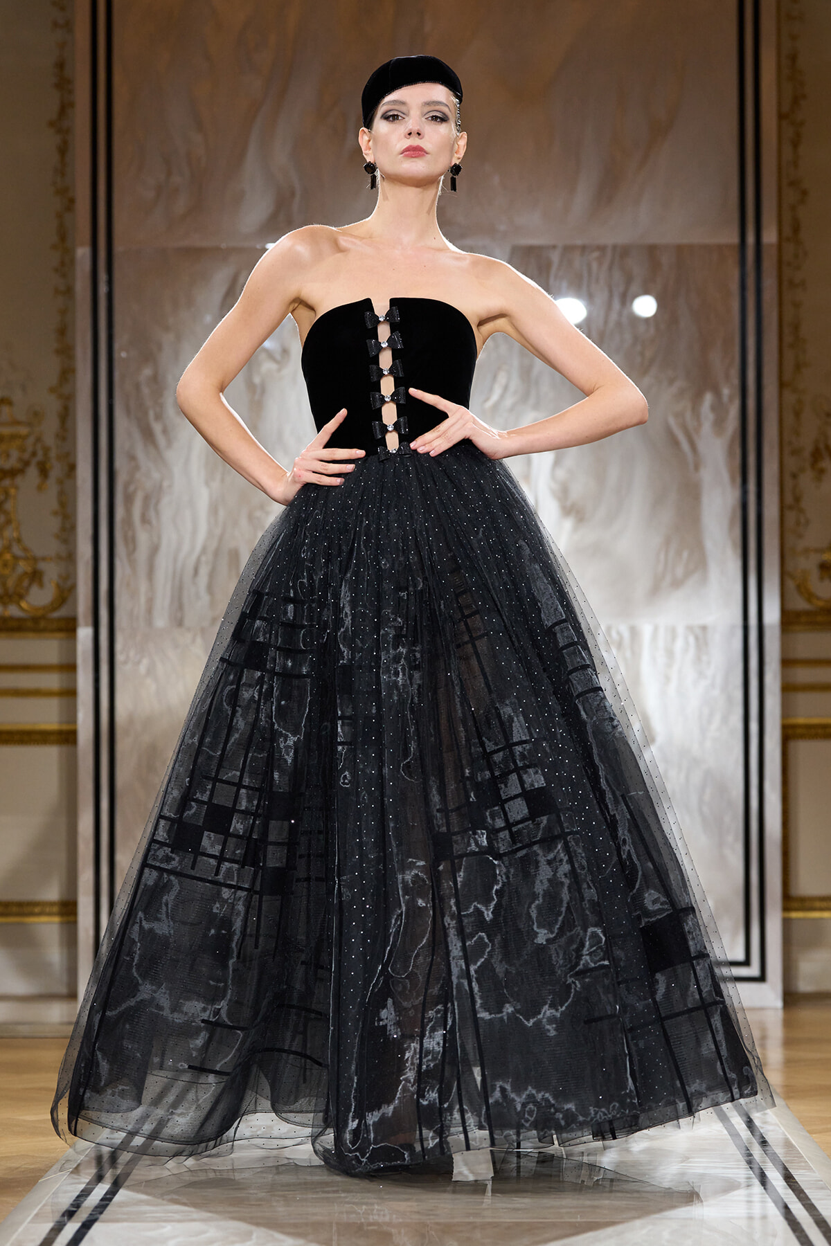 Model on a runway wearing a strapless black ball gown with a laced front bodice and a voluminous dotted tulle skirt, complemented by a black cap and statement earrings.