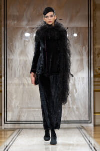 Model on a runway wearing a black velvet pinstripe suit with a dramatic feathered scarf and matching headpiece, carrying a small clutch.