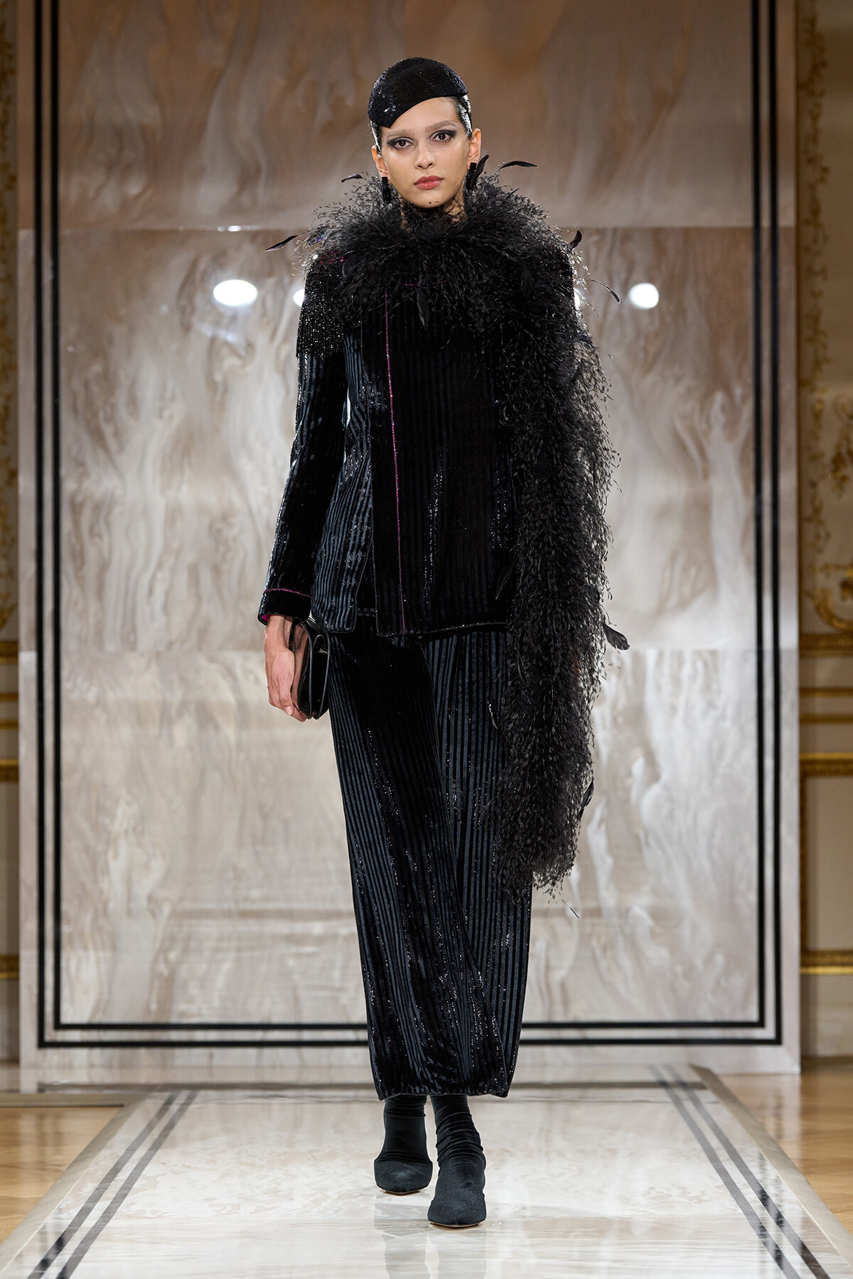 Model on a runway wearing a black velvet pinstripe suit with a dramatic feathered scarf and matching headpiece, carrying a small clutch.