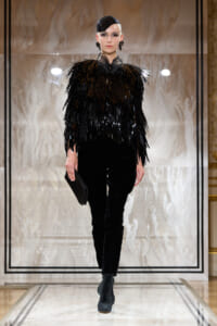 Model walks a runway in an all-black outfit: a fringed, sequined top with velvet pants and heeled ankle boots, carrying a clutch.