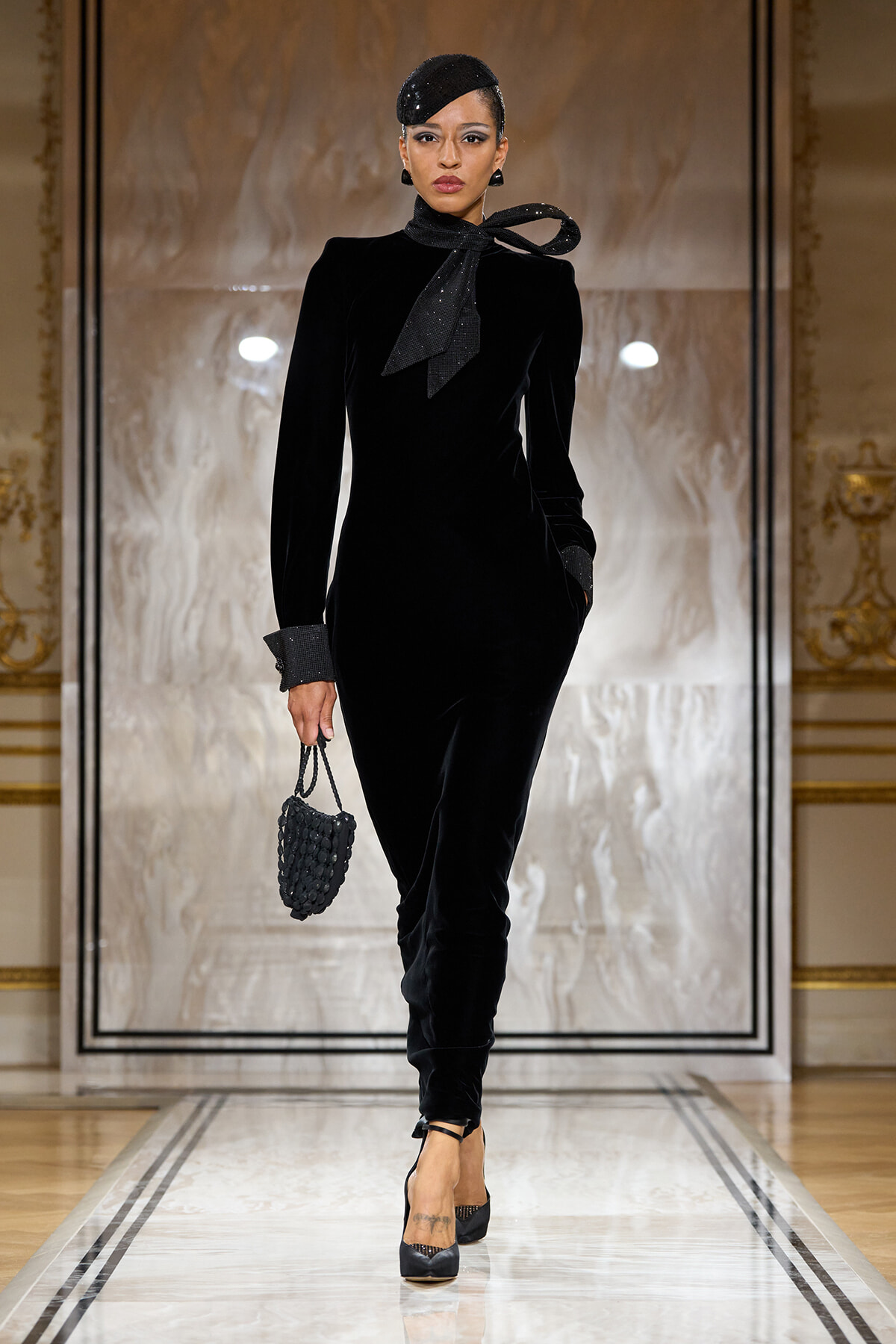 Model in a black velvet dress with a tied neck scarf, wearing a small black woven handbag, on a fashion runway.