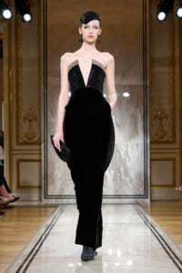 Model walks the runway in a floor-length black velvet gown with a plunging beaded bodice and crystal accents at the shoulders.
