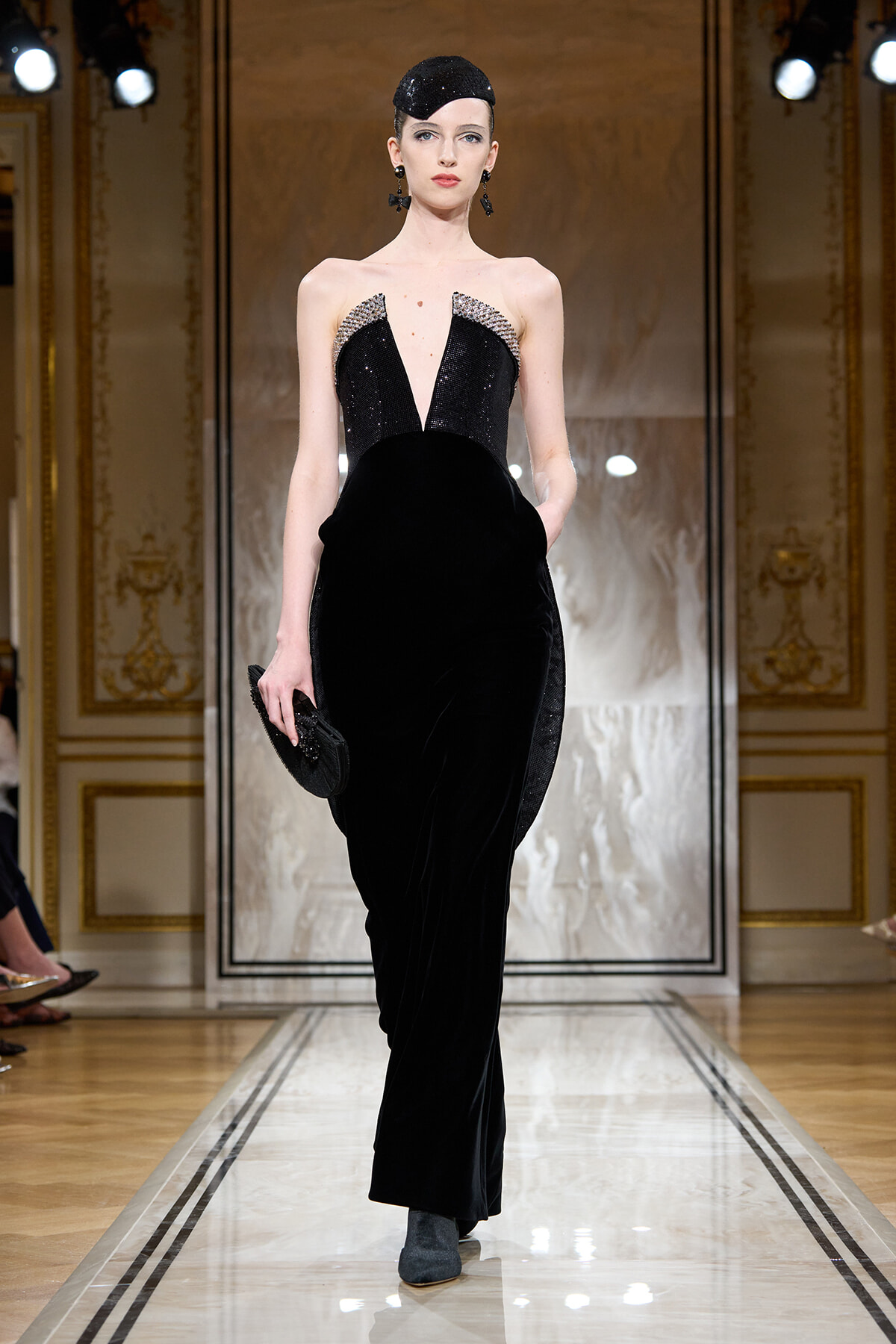 Model walks the runway in a floor-length black velvet gown with a plunging beaded bodice and crystal accents at the shoulders.