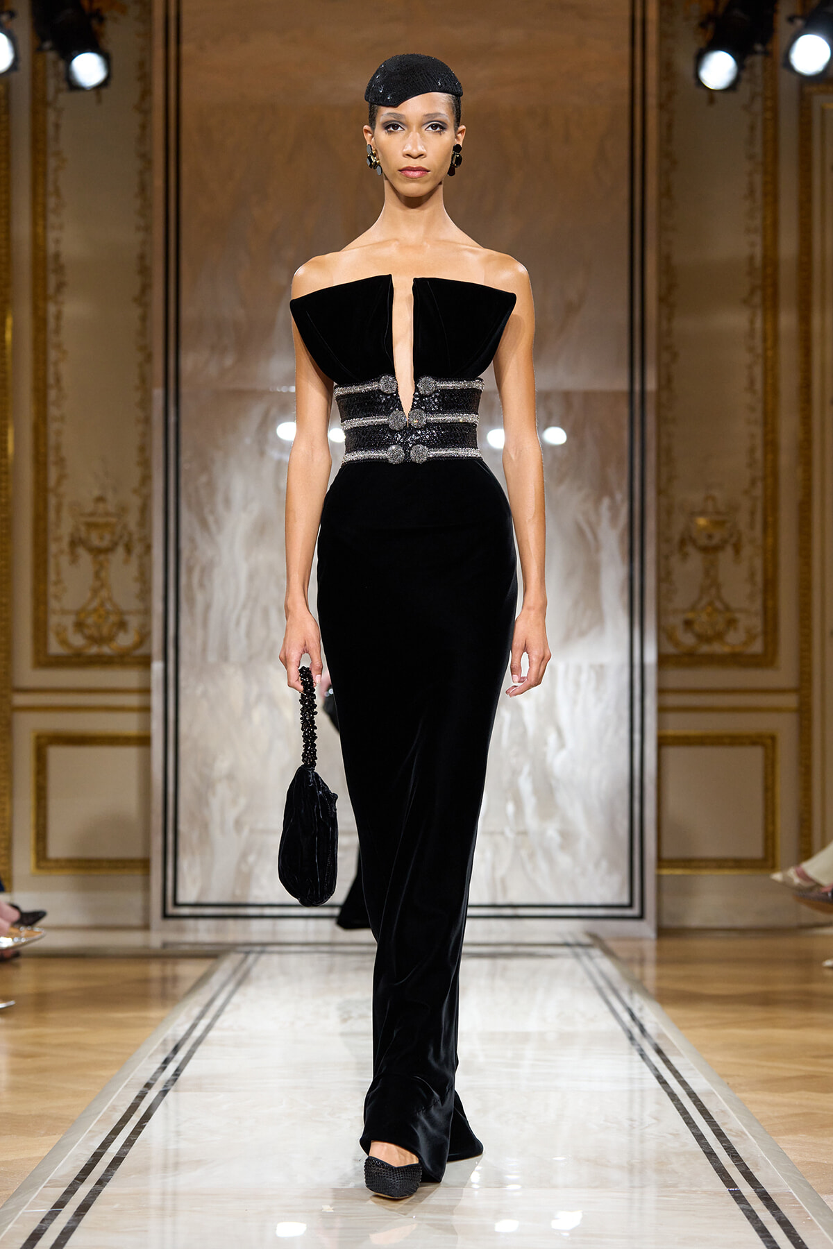 Model walking runway in a black velvet gown with a deep V neckline and jeweled waist belt, holding a black clutch.