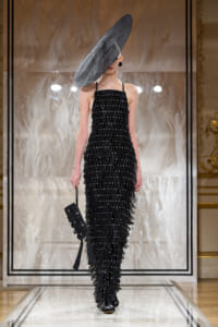 Model on a runway wearing a floor-length black beaded fringe gown with a large tilted circular hat, carrying a black clutch.