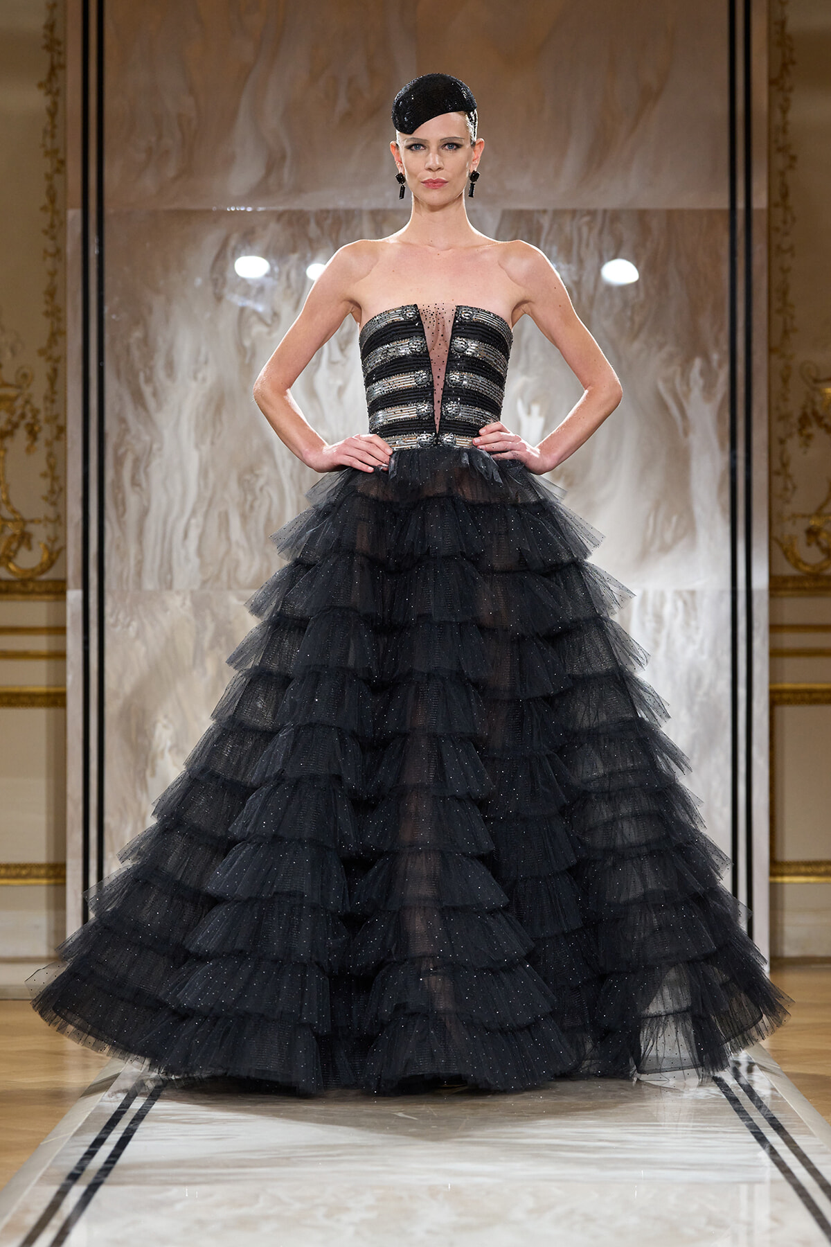 Model on a runway wearing a black strapless sequined corset with a voluminous layered tulle ball gown.