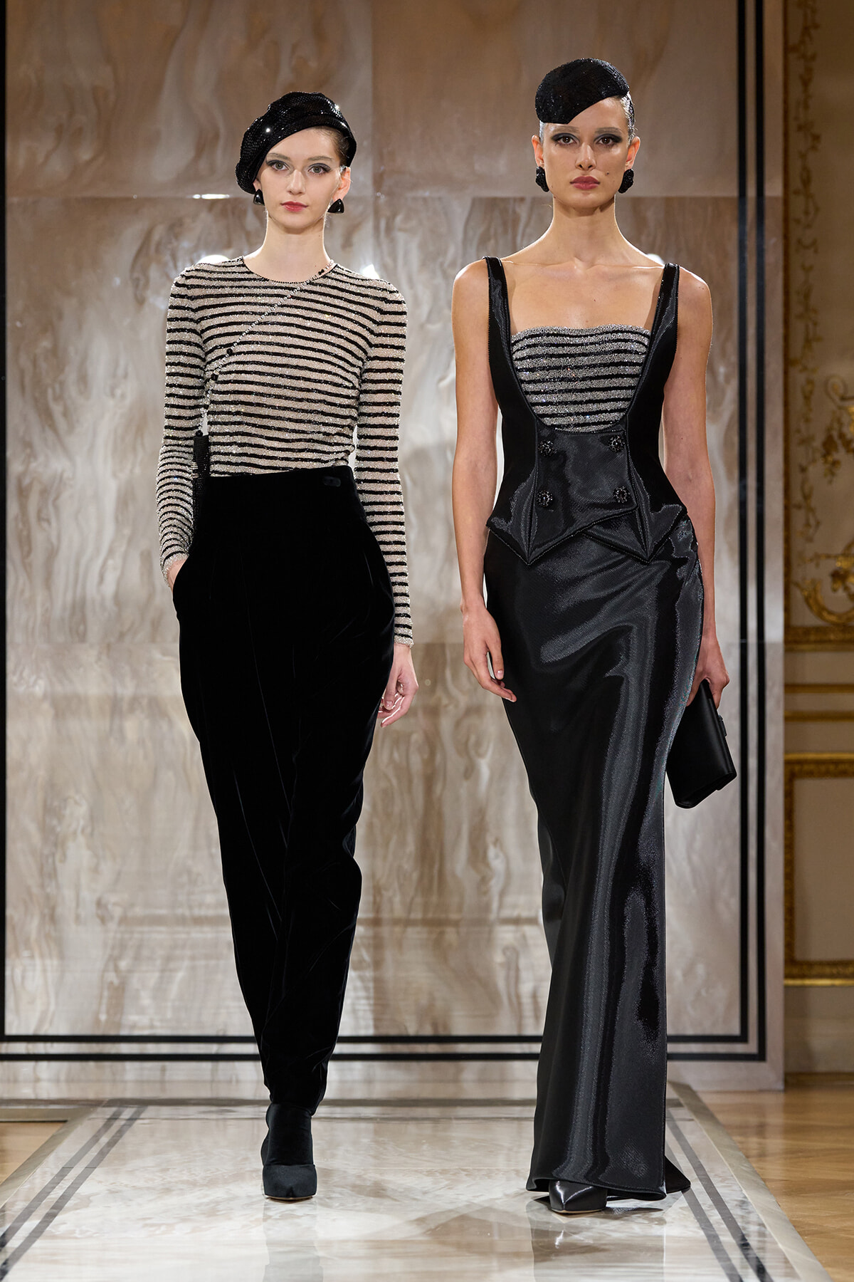 Two models walk a runway in black and silver evening wear—left in velvet pants with a striped top, right in a strapless, metallic-bodice gown.