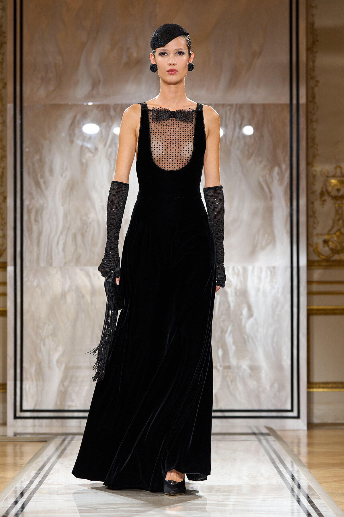 Model on a runway wearing a floor-length black velvet gown with a sheer, polka-dot bodice, long beaded gloves, and a small black fascinator.