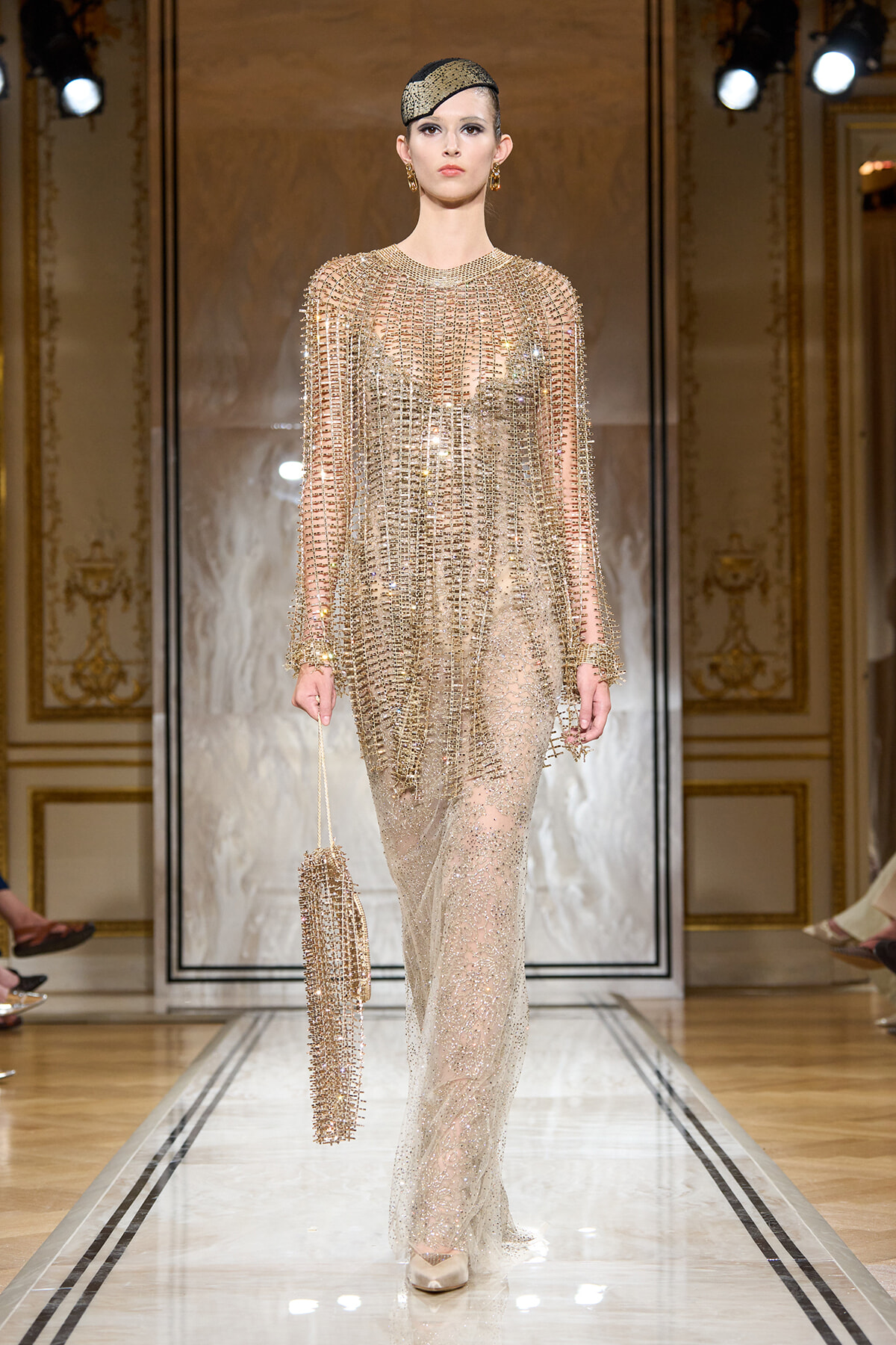 Model walking a couture runway in a gold, beaded sheer gown with long sleeves, paired with a small beaded clutch and a gold headpiece in an ornate, chandelier-lit venue.