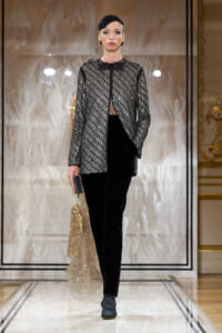 Runway model wearing a silver-embellished jacket over a sheer top and black velvet pants, clutch in hand, on a marble catwalk.