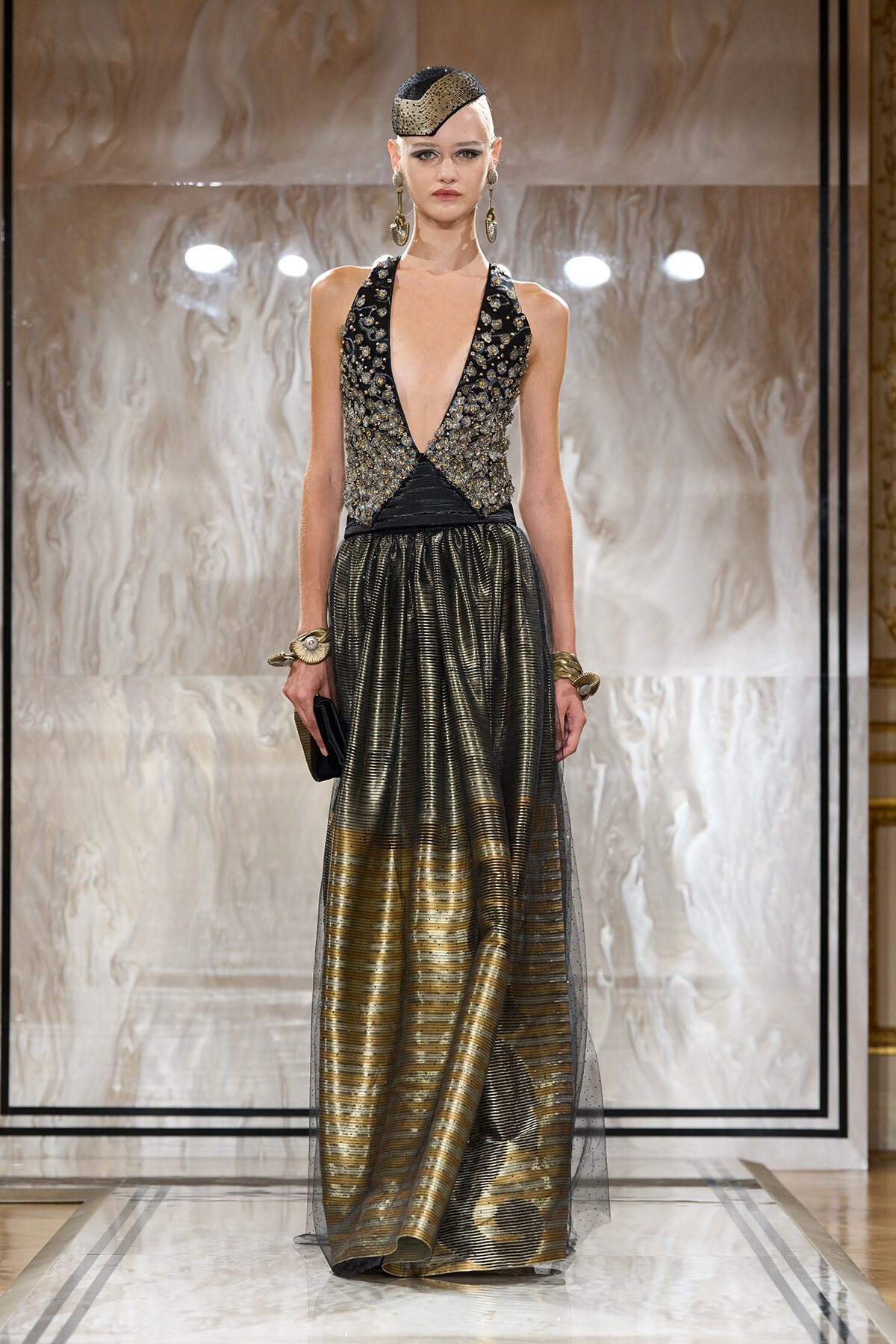 Model on a runway wearing a black-and-gold sequined halter bodice and a metallic striped skirt with sheer overlay, gold headpiece and jewelry.