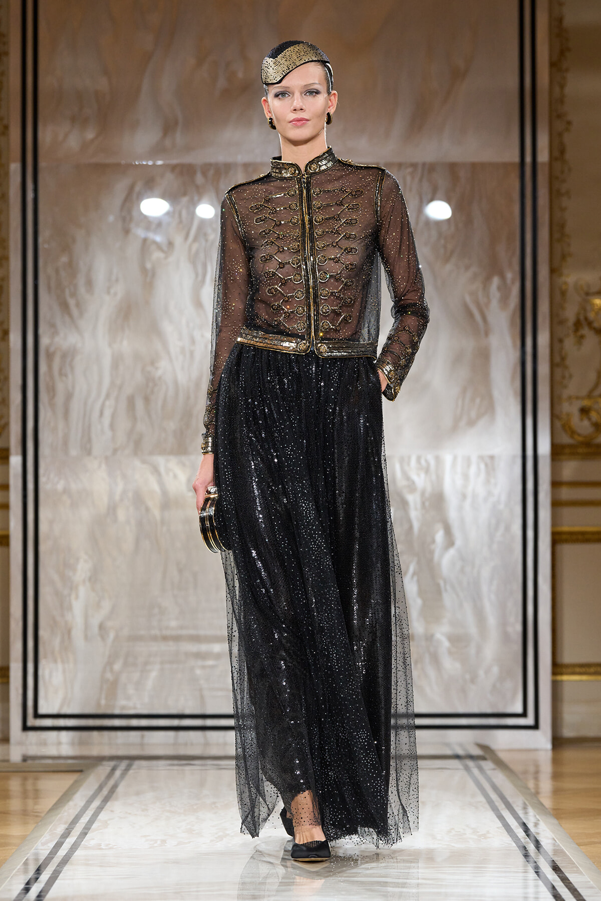 Model walking the runway in a sheer black embellished jacket with gold trim over a glittering black maxi skirt, accessorized with a gold-sequined cap and drop earrings, and holding a small gold clutch.