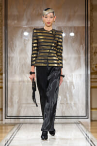 Model walks a runway in a metallic gold-and-black striped structured jacket with a black satin belt, paired with glossy black wide-leg pants and black boots, accessorized with a beaded headpiece and gold earrings.