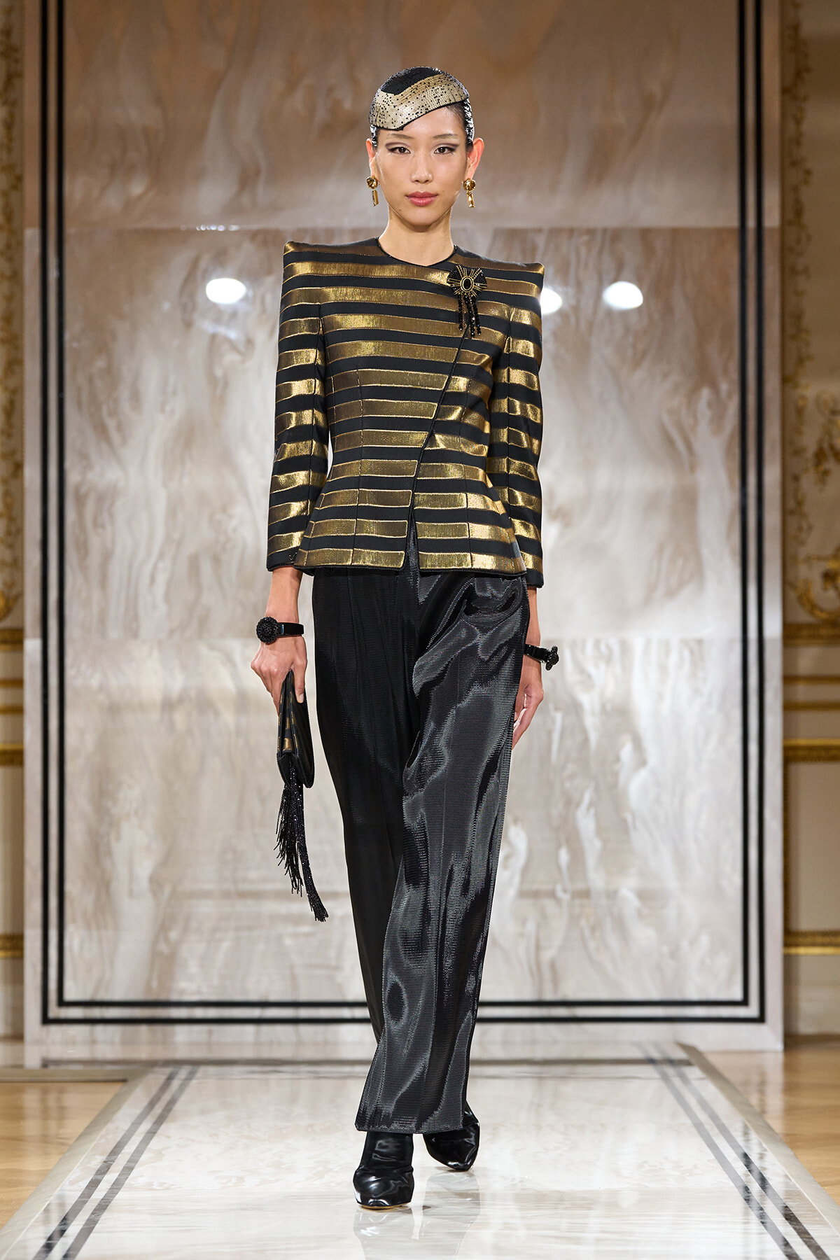 Model walks a runway in a metallic gold-and-black striped structured jacket with a black satin belt, paired with glossy black wide-leg pants and black boots, accessorized with a beaded headpiece and gold earrings.