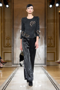Runway model wearing a black embroidered jacket with gold floral details and two-tone velvet trousers, carrying a small clutch and wearing a sequined cap.