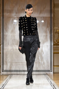Runway look: model in a black velvet embellished jacket with metallic buttons, sheer gloves, and glossy satin trousers.
